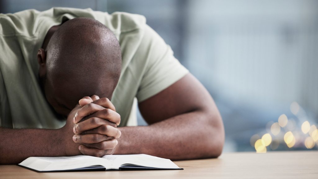 A close-up of a man leaning over an open Bible with his hands clasped together, practicing a peaceful moment of deep prayer and scripture study while learning how to grow in faith.